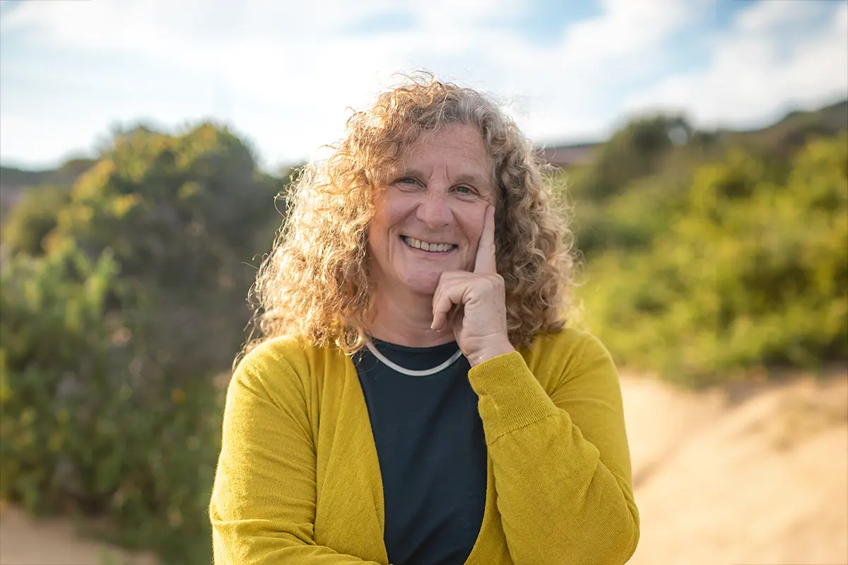 Gabriele Mann (geborene Reichart) trägt einen senffarbenen Cardigan und steht in der Natur. Dabei lächelt sie in die Kamera während sie mit der linken Hand ihr Gesicht aufstützt.