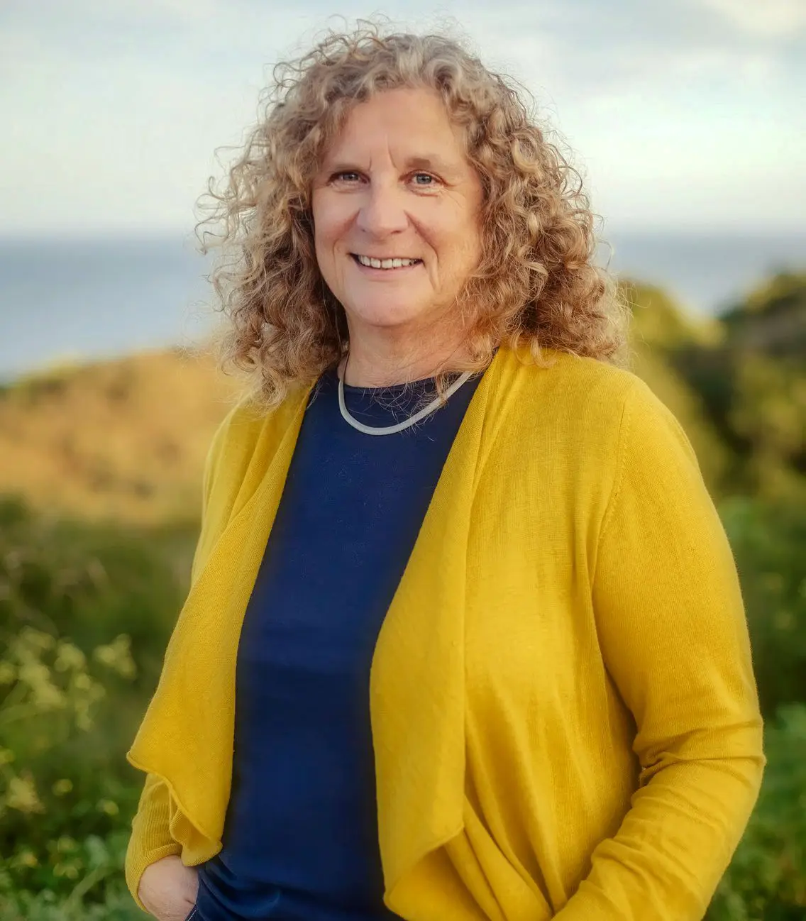 Gabriele Mann (geborene Reichart) trägt einen senffarbenen Cardigan und lächelt in der Natur vor dem Meer stehend für ihr Über mich Foto in die Kamera.