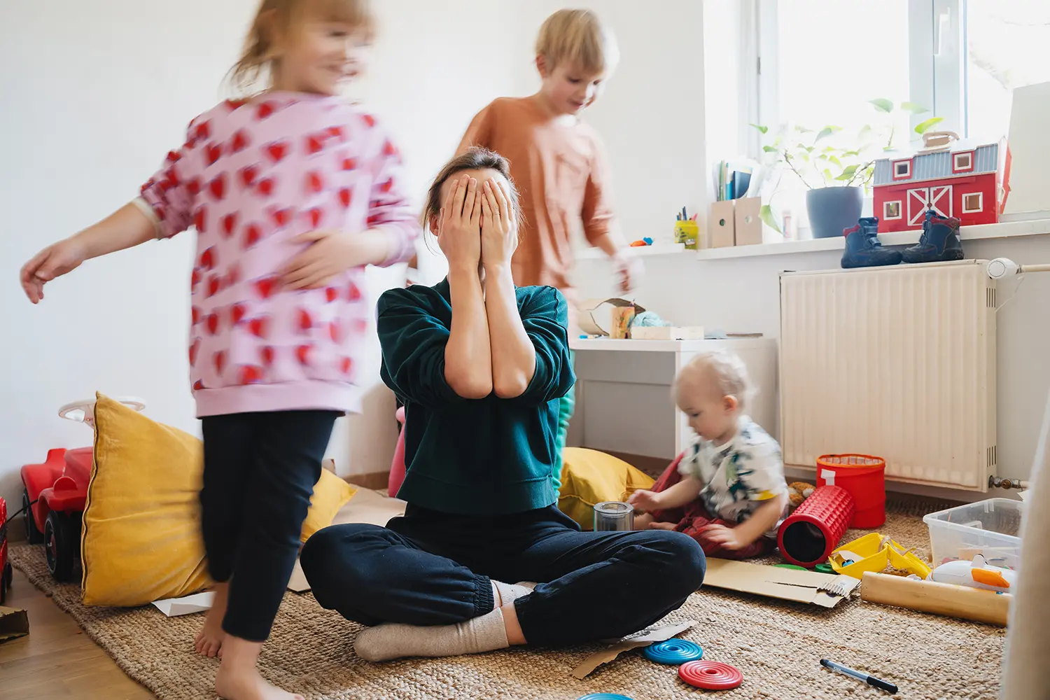 Eine überforderte Mutter sitzt im Spielzimmer zwischen ihren drei tobenden Kindern als Symbol für Grenzen setzen bei Kindern.