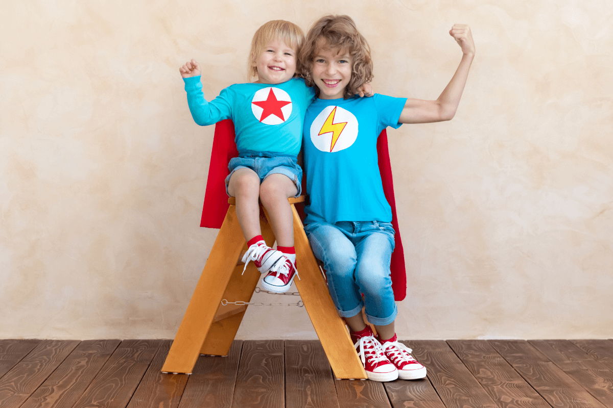 Zwei Kinder - ein älteres ca. 8 Jahre alt und ein kleineres, ca. 2 Jahre alt sitzen auf einer kleinen Holzleiter. Beide lächeln fröhlich und halten je einen Arm mit einer Faust geballt zur Seite. Es entsteht ein Bild der Stärke.