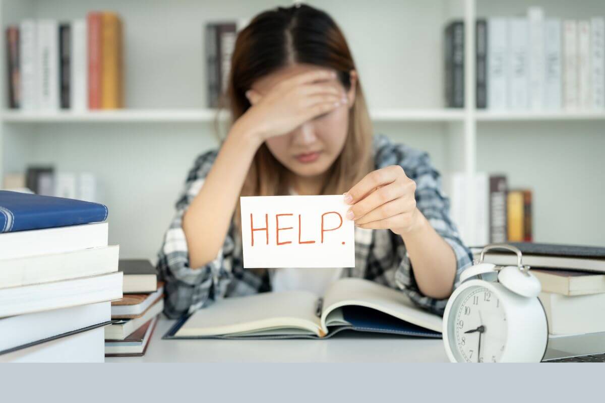 Erschöpfte Mutter sitzt am Tisch über Büchern und hält ein Schild "Help" in der Hand. Sie ist müde, weil sie den ganzen Tag ihr Kind an alles erinnert, weil es an nichts selbst denkt.