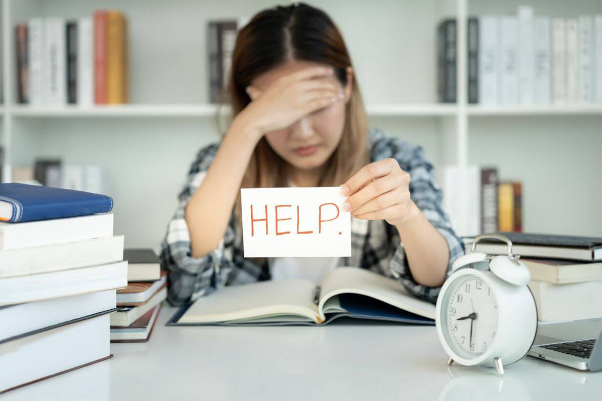 Erschöpfte Mutter sitzt am Tisch über Büchern und hält ein Schild "Help" in der Hand. Sie ist müde, weil sie den ganzen Tag ihr Kind an alles erinnert, weil es an nichts selbst denkt.