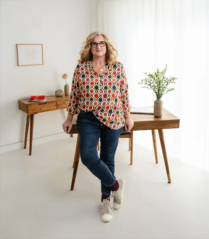 Gabriele Reichart von Die Eltern Akademie auf ihrer Herzensmission: Liebevolle Erziehung leicht gemacht. Sie steht entspannt vor und stützt sich auf ihren Schreibtisch, während sie lächelnd in die Kamera blickt.